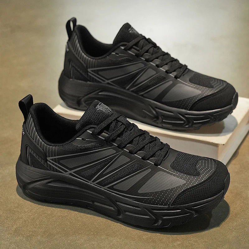 A pair of black athletic sneakers with textured mesh and synthetic overlays, displayed on a neutral concrete floor with one shoe resting on a white box.