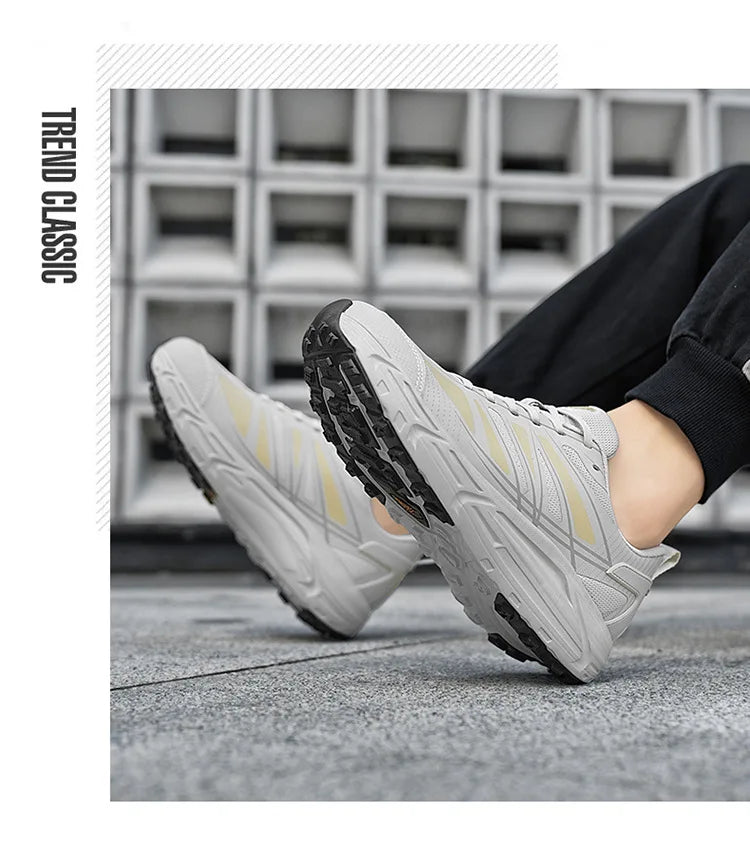 A pair of light gray athletic sneakers with pale yellow stripes and black soles, shown being worn with black pants. The background is a modern gray concrete block wall and textured pavement.