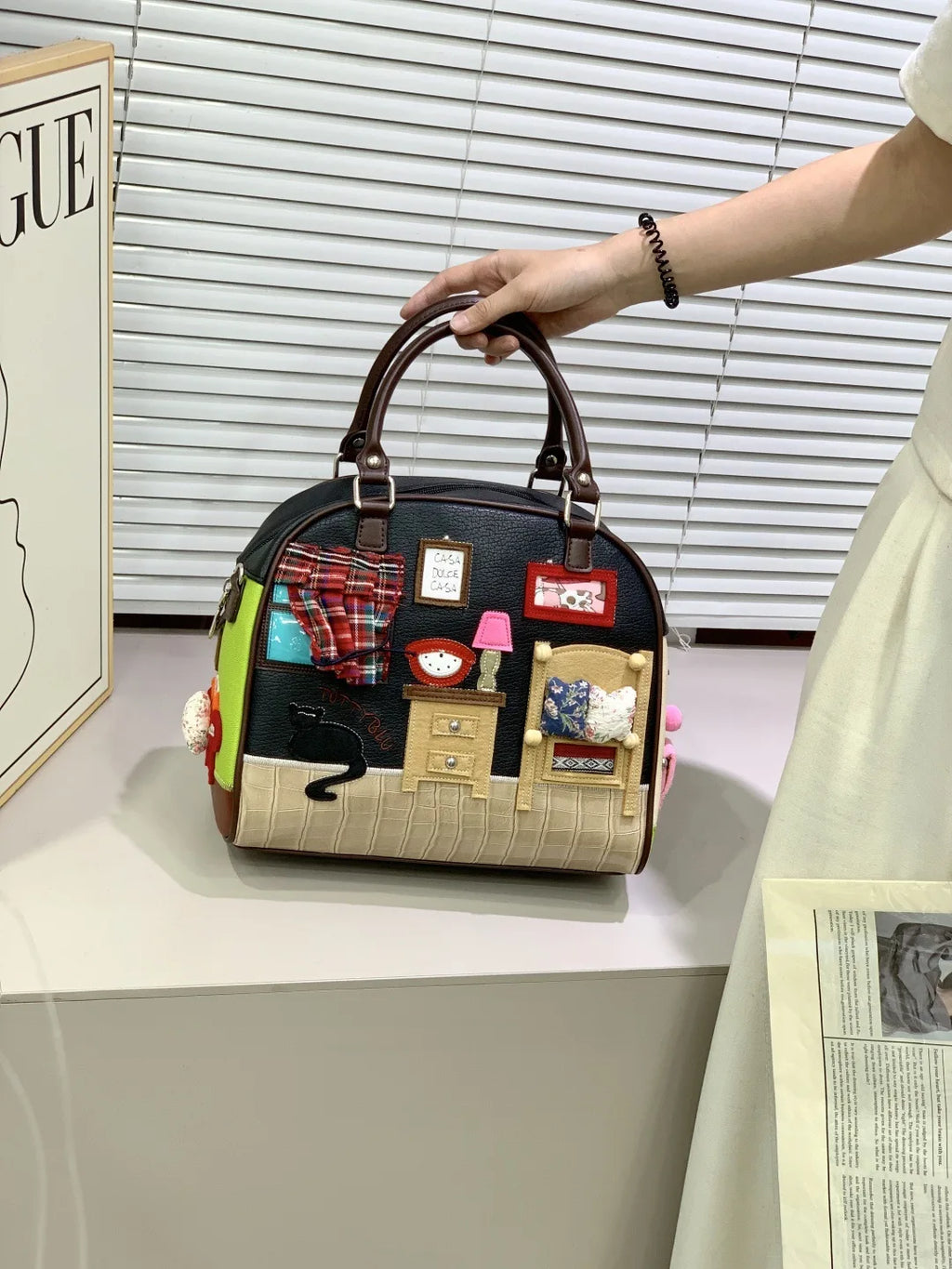Handbag with decorative patches held by a person, with a magazine and window blinds in the background.