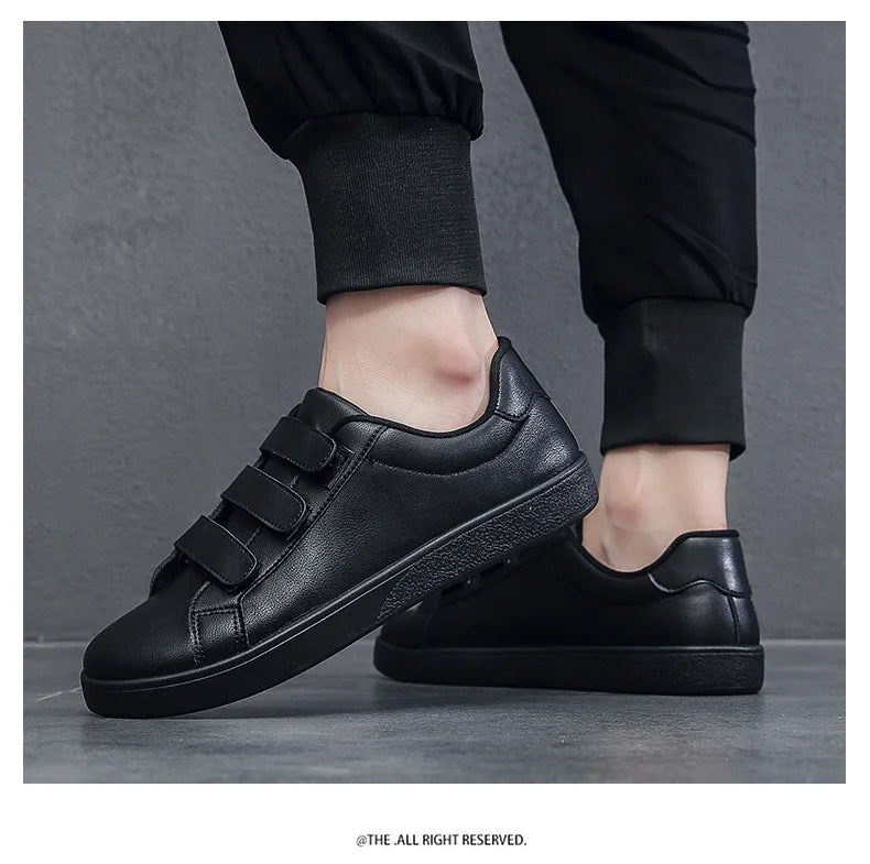 Black sneakers with velcro straps worn by a person on a gray background