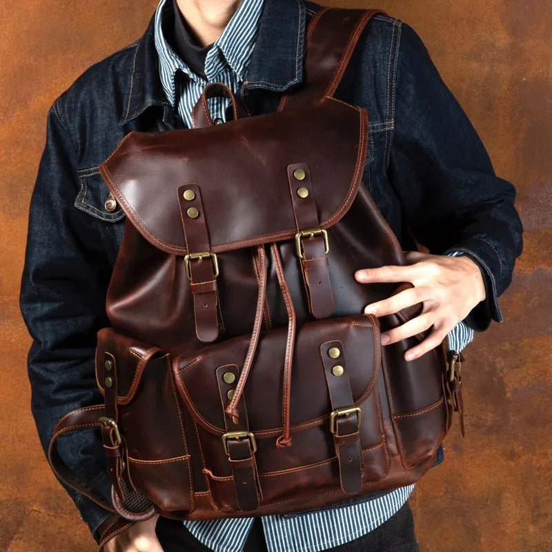 A dark brown leather backpack with brass hardware, worn by a person wearing a blue denim jacket over a striped shirt, set against a textured brown background.