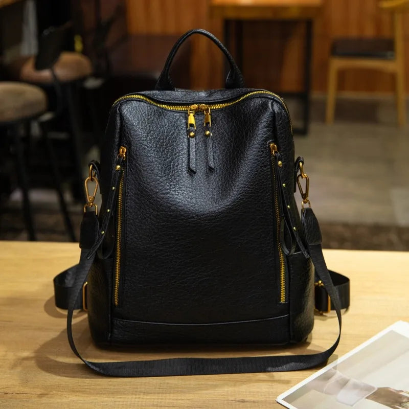Black pebbled leather backpack with gold-tone zippers and hardware, displayed on a wooden table in a cafe setting.