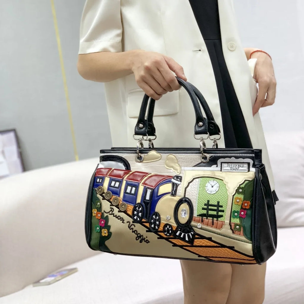 A person in a cream blazer holds a structured handbag with black handles. The bag has a beige base with a colorful train illustration and the words 'Susan Maggie' on the front.