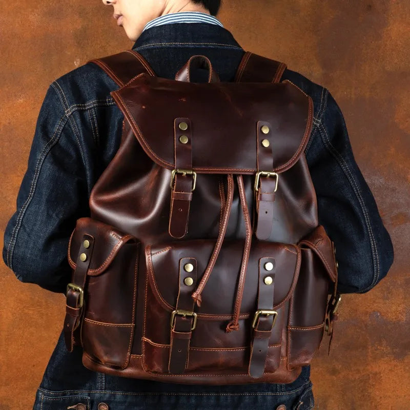 A person viewed from behind wearing a dark brown leather backpack with multiple front pockets and brass-tone hardware, over a dark denim jacket against a textured brown background.