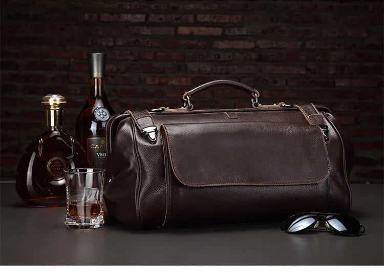 Brown leather duffel bag with a bottle of Cognac, glass, and sunglasses on a dark background