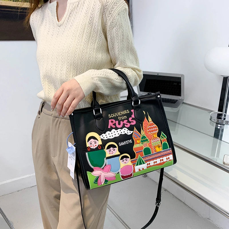A person wearing a cream cable-knit sweater and beige pants holds a black handbag decorated with colorful Russian-themed illustrations, including nesting dolls, the text 'SOUVENIRS from RUSSIA', and a pink bow.