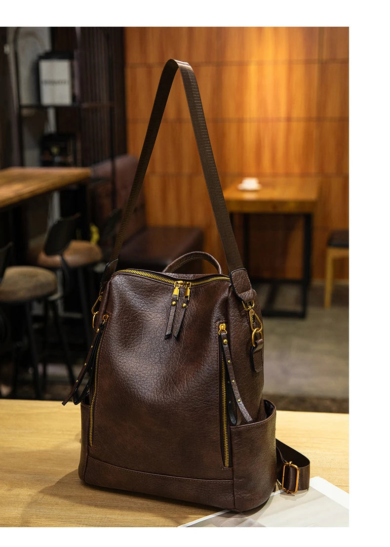 A dark brown textured leather shoulder bag with gold-tone hardware and multiple zippered pockets, displayed on a wooden table in a warm, modern cafe setting.
