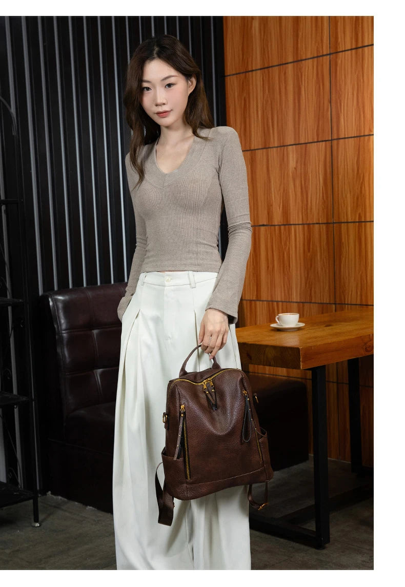 A woman stands indoors holding a brown leather backpack. She is wearing a fitted taupe long-sleeve V-neck top and white wide-leg pants.