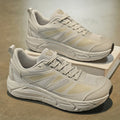A pair of chunky low-top athletic sneakers in a light beige and off-white colorway, displayed on a neutral textured floor with one shoe resting on a small white box.