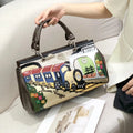 A hand holds a structured handbag with a brown leather top handle and trim, featuring a colorful 3D train illustration on the front. The handbag is shown on a light beige sofa next to a person wearing a cream-colored blazer.