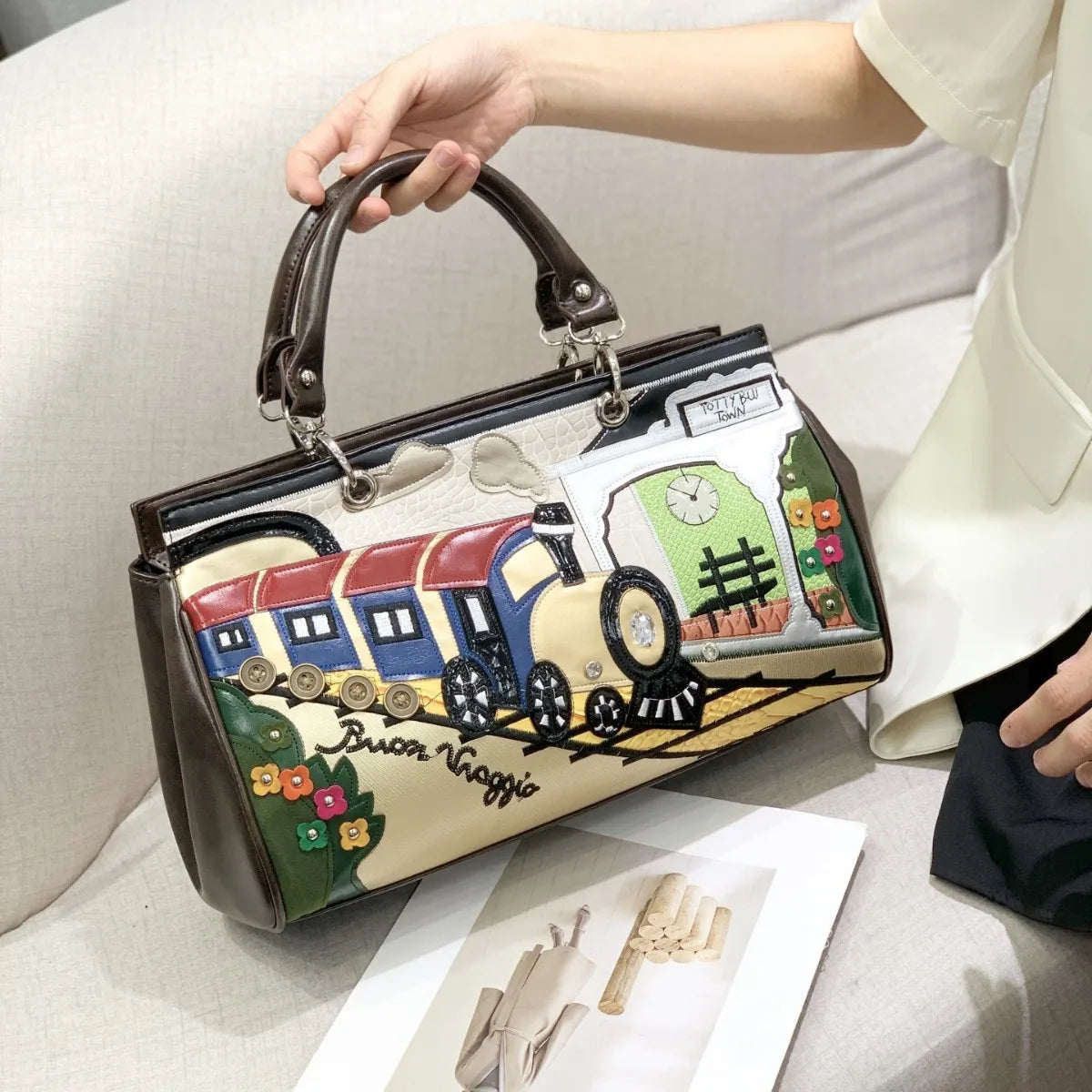 A hand holds a structured handbag with a brown leather top handle and trim, featuring a colorful 3D train illustration on the front. The handbag is shown on a light beige sofa next to a person wearing a cream-colored blazer.