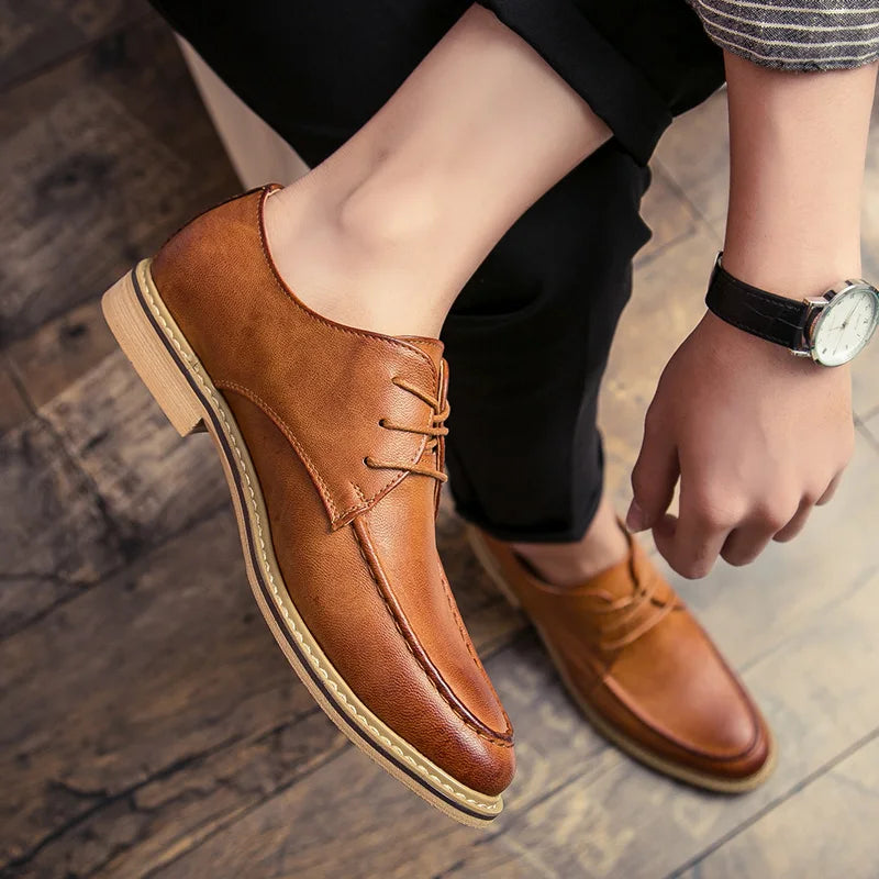 A person wearing a pair of brown leather lace-up dress shoes with low block heels, shown from the knees down while sitting on a wooden floor. The person is wearing black trousers and a striped shirt, and has a black leather-strap wristwatch on.