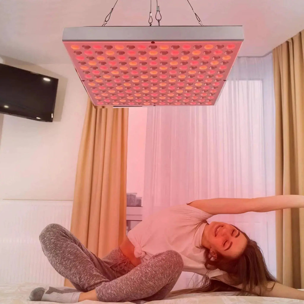 Person lying on a bed under a red light therapy panel in a room with curtains.