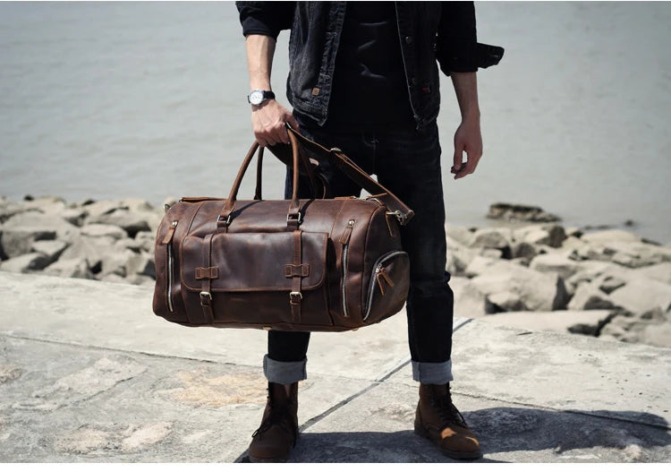 Person holding a brown leather duffel bag by a rocky shoreline