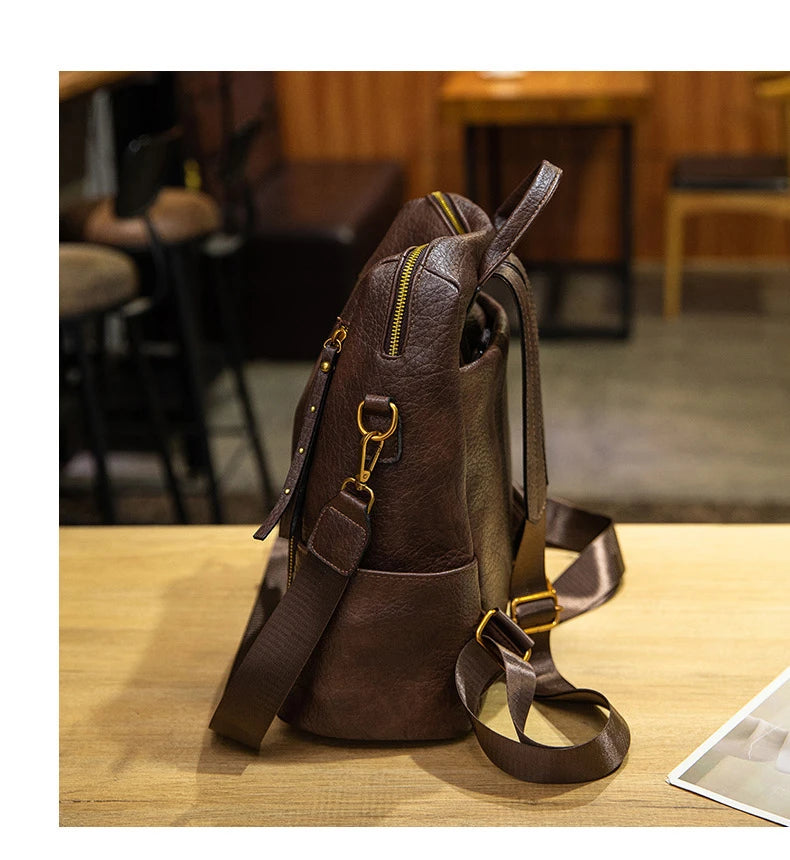 A dark brown faux-leather backpack with gold-tone hardware and a matching shoulder strap, displayed on a light wooden table in a cafe setting.