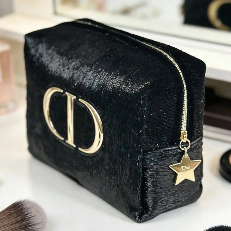 A black textured cosmetic pouch with a gold-tone 'CD' logo and a dangling star charm, photographed on a light tabletop with blurred makeup items in the background.
