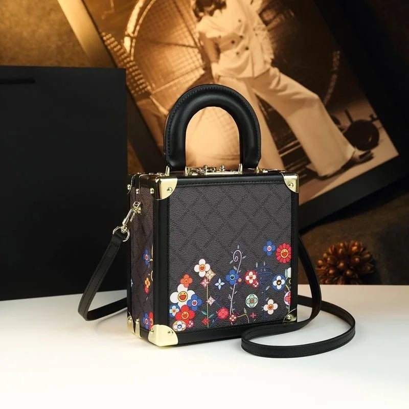 A structured black handbag with a floral print on the front, gold-tone hardware, a top handle, and a detachable shoulder strap, displayed on a white surface with a black box and framed photo in the background.