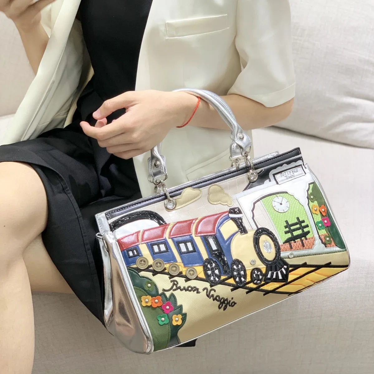 A person seated on a sofa holds a shiny silver handbag with a colorful train illustration and the text 'Brian Maggie' on the front.