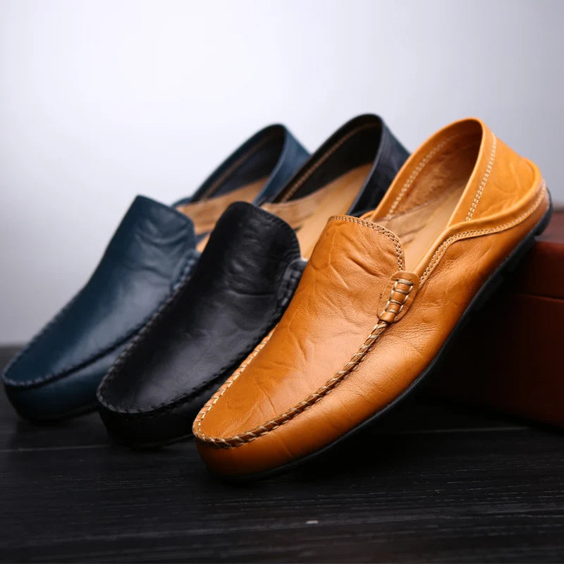 Three leather slip-on loafers arranged on a dark surface against a light gray background: one tan/cognac, one black, and one dark navy. The tan shoe is in front and most prominent, with the other two partially visible behind it.