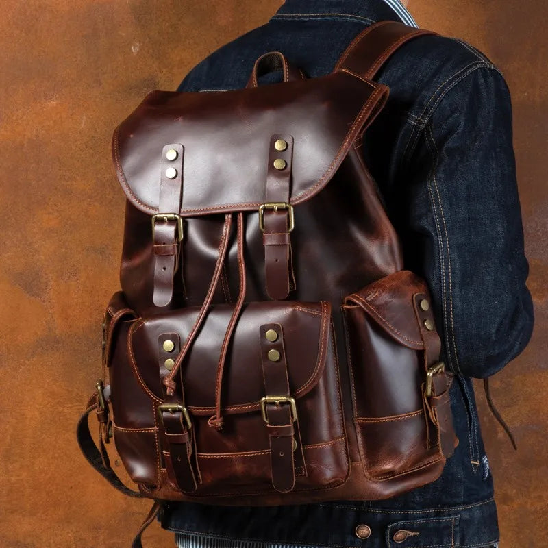 A dark brown leather backpack with brass hardware, shown from the back on a person wearing a dark denim jacket against a textured brown background.