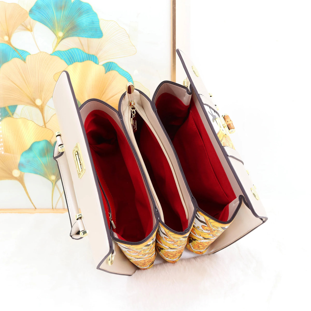 A cream-colored shoulder bag with gold-tone hardware, shown open to display its deep red interior lining and gold patterned fabric trim. The bag is positioned in front of a decorative framed print featuring gold and teal ginkgo leaves on a white background.