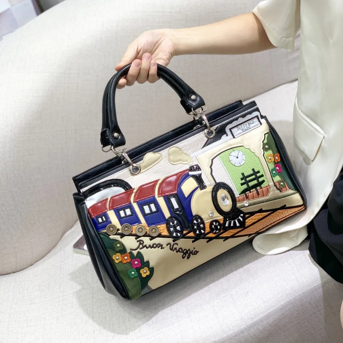 A hand holds a structured handbag with a black handle, featuring a colorful printed design of a vintage train on a cream background. The person holding the bag is wearing a white blouse and dark skirt, sitting on a light-colored fabric sofa.