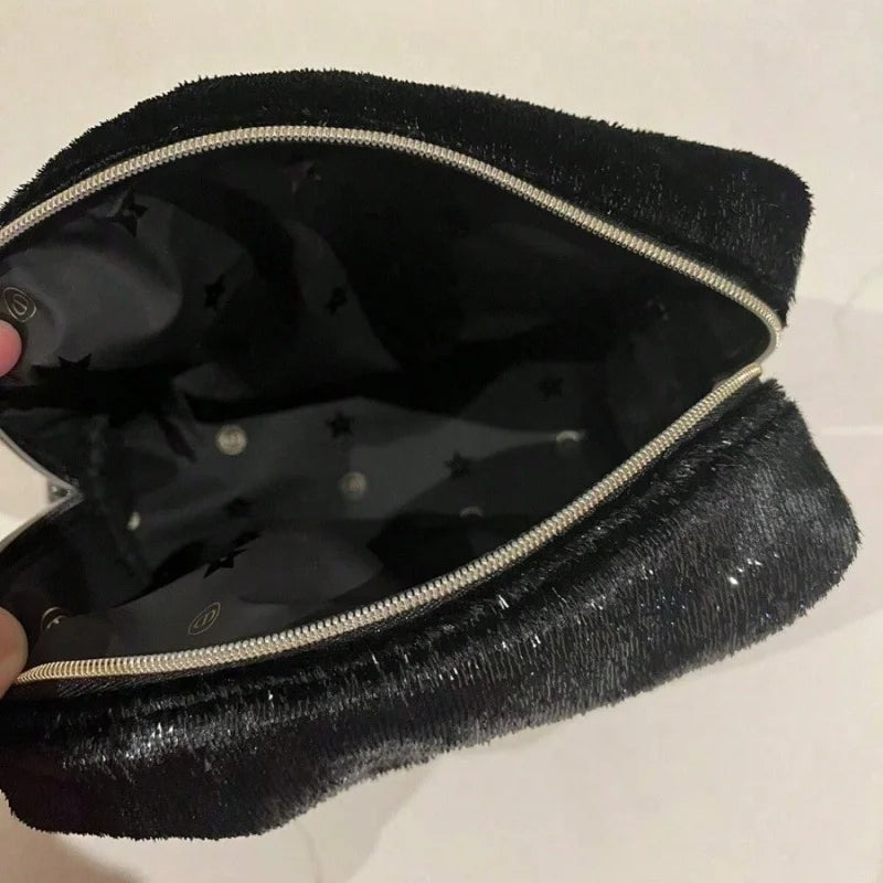 A black plush or velour zippered pouch shown open, revealing a black star-patterned interior and a silver zipper. A hand is holding the left edge of the pouch against a plain light background.