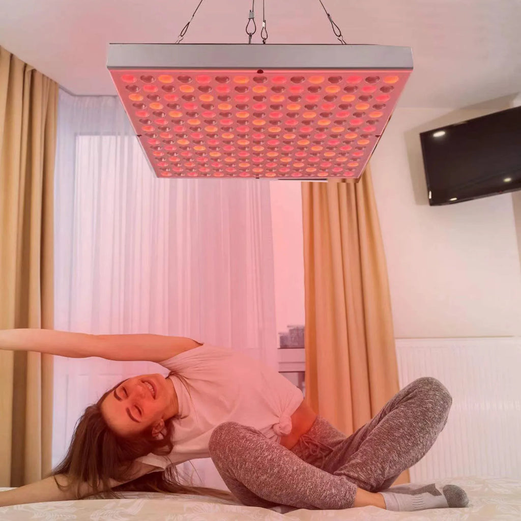 Person lying on a bed under a red light therapy panel in a room with curtains and a TV.