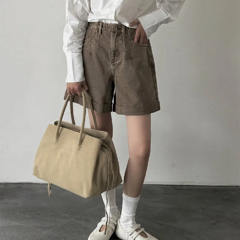 Person wearing brown shorts and white shoes, holding a beige bag against a plain background