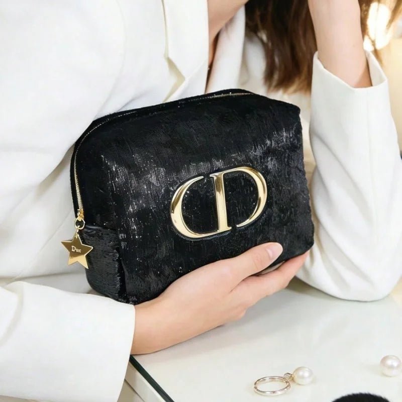 A black textured cosmetic pouch with a large gold-tone 'CD' logo and a small gold star charm, held by a person wearing a white blazer.