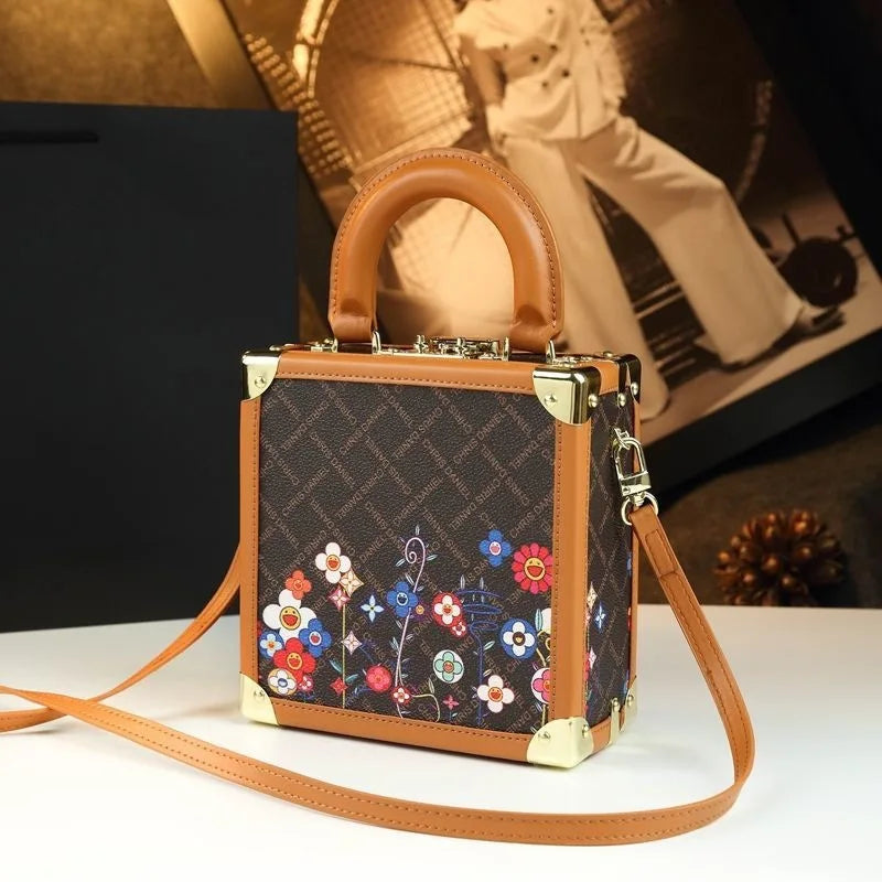 A structured mini handbag with a brown monogram canvas body, colorful floral embroidery on the lower front, tan leather trim and top handle, and gold-tone hardware, shown on a tabletop with a matching shoulder strap.