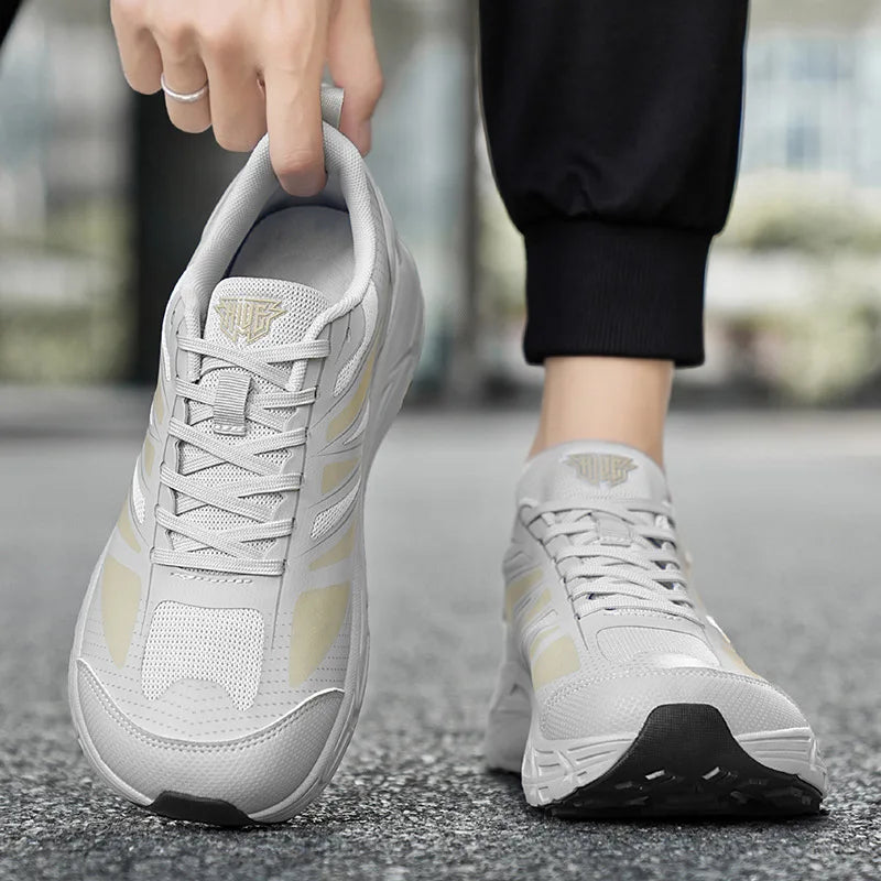 A person wearing light gray and beige athletic sneakers, with one shoe being held up by a hand. The shoes feature a mesh upper with light gray panels, beige accents, and a black outsole, set against a blurred outdoor background.