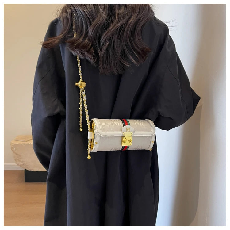 A person wearing a black oversized coat carries a small structured shoulder bag with a gold chain strap. The bag is light beige with an embossed monogram pattern and a red and green web stripe detail on the front, with gold hardware.