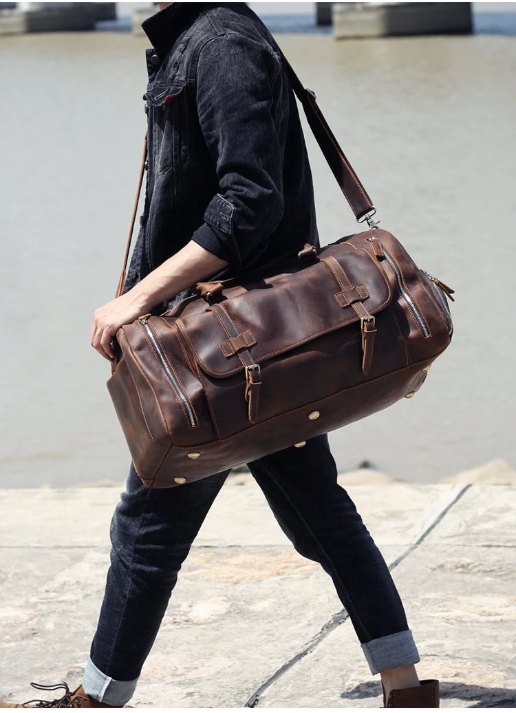 Person holding a large brown leather duffel bag outdoors.