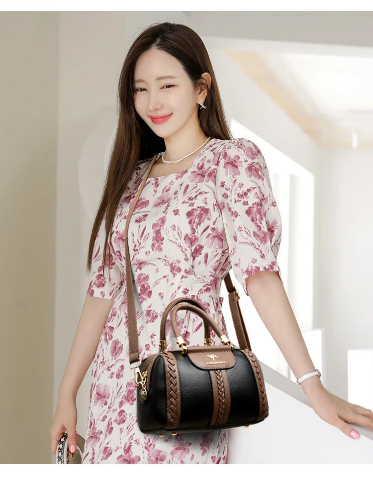 A woman wearing a pink and white floral dress carries a black and brown leather handbag with braided trim and gold-tone hardware in a bright indoor setting.
