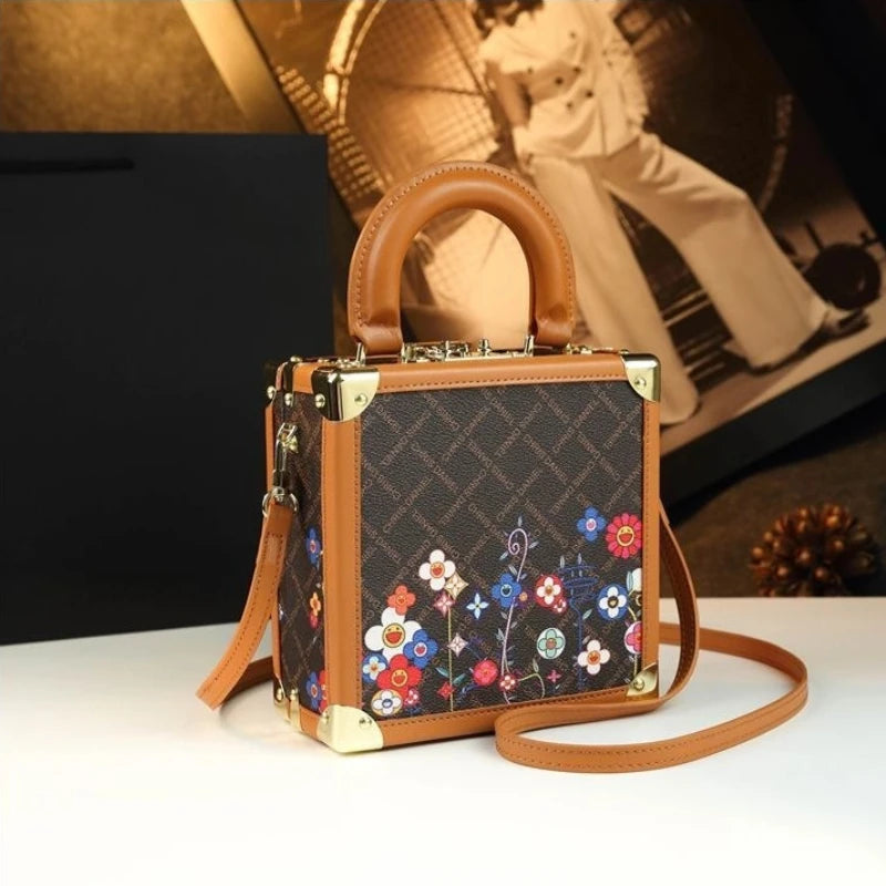 A structured handbag with a brown leather top handle and shoulder strap, featuring a dark brown monogram patterned body with colorful floral embroidery along the bottom, and gold-tone metal corner protectors, displayed on a white surface with a warm-toned background.