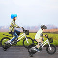 Two children riding white balance bikes with training wheels on a road with a grassy field and sky in the background.