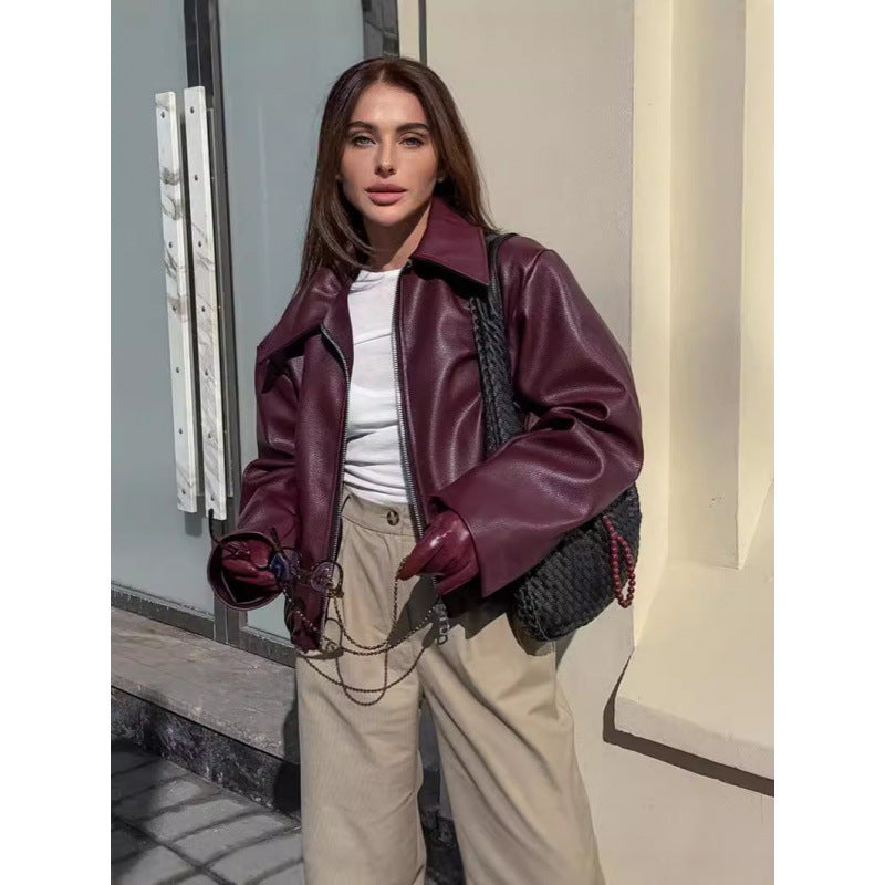 Woman wearing a burgundy leather jacket and beige pants, standing against a building exterior.