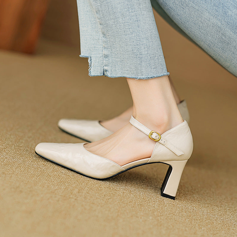 A close-up of a person wearing light beige pointed-toe heeled pumps with ankle straps and gold buckle details, paired with light blue jeans, standing on a beige carpeted floor.
