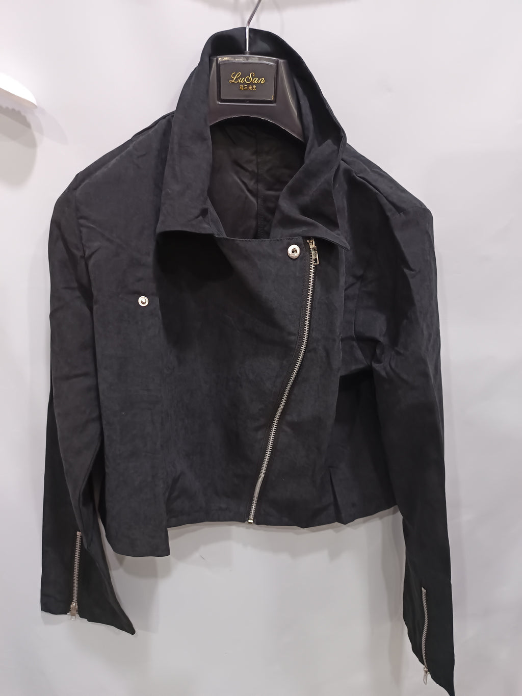 A black cropped moto jacket with an asymmetrical zipper, displayed on a hanger against a plain white wall. The brand label on the collar reads 'LeLieu New York'.