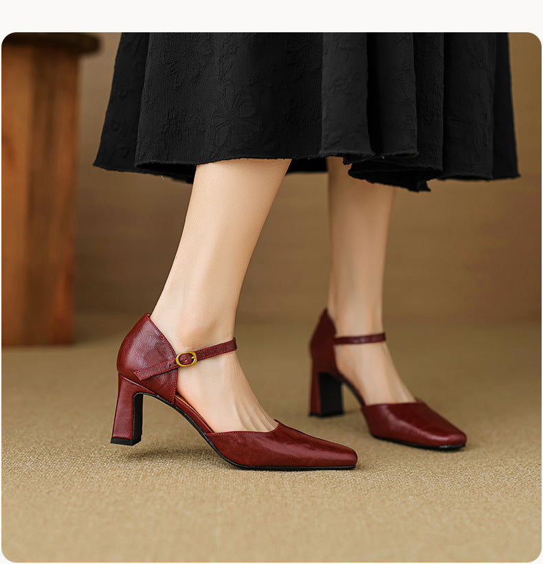 A person wearing a black skirt and glossy dark red Mary Jane-style block-heel shoes with ankle straps and small gold-tone buckle details, standing on a beige floor in an indoor setting.