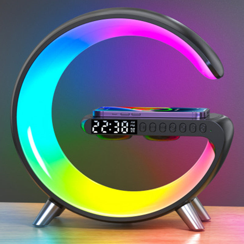 A modern C-shaped LED desk lamp with a built-in smartphone charging dock and digital clock display, shown illuminated with a rainbow gradient on a tabletop against a dark gradient background.