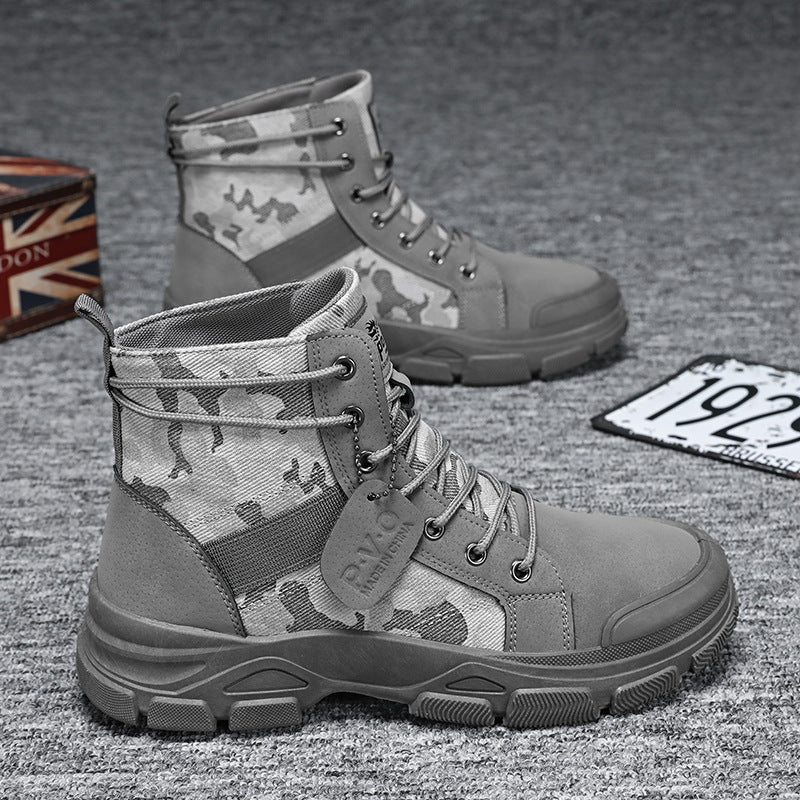 A pair of gray and camouflage mid-calf lace-up boots displayed on a textured gray surface, with a small Union Jack box and a license plate prop in the background.